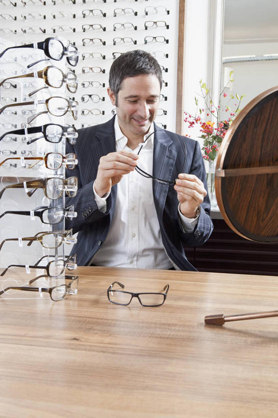 西方人,一个人,商店,站,笑,微笑,露齿笑,竖图,室内,白天,正面,西装,桌面,桌子,欧洲,欧洲人,仅一个男性,仅一个人,仅一个中年男性,握,套装,正装,朦胧,模糊,木制,眼镜,拿,拿着,注视,职业装,商铺,货架,架子,木制品,观察,看,站着,握着,观看,验光师,察看,关注,陈列架,近视眼镜,眼镜店,超市货架,市肆,商号,男人,男性,中年男性,白种人,站立,半身,彩图,店铺
